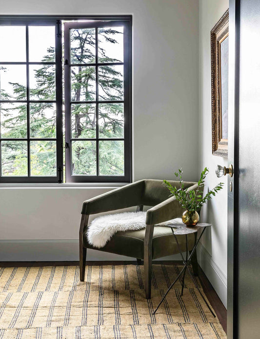| The Lyssa olive velvet accent chair sits in the corner of a room with a striped rug and a white sheepskin blanket thrown atop it.