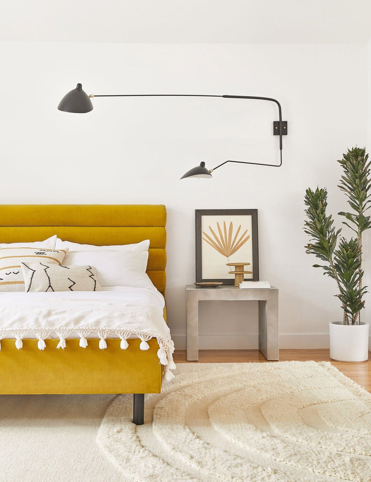 | The Gin indoor and outdoor concrete side table sits in a bedroom with a golden linen bed, a floral wall art, and a white plush rug