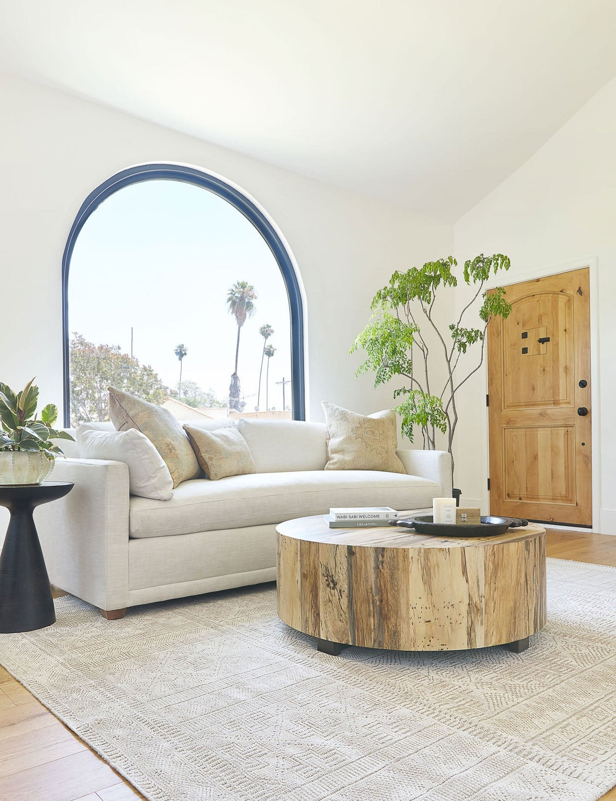 | The Boni round coffee table sits in a living room with a large arched window, an ivory rug, and an ivory sofa