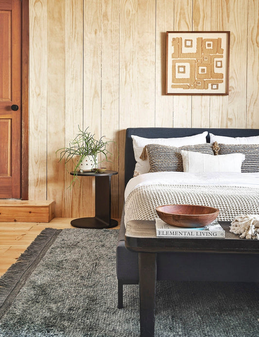 | The Illy metal geometric side table sits in a bedroom with a gray linen bed, a gray plush rug, and a light wood paneled wall