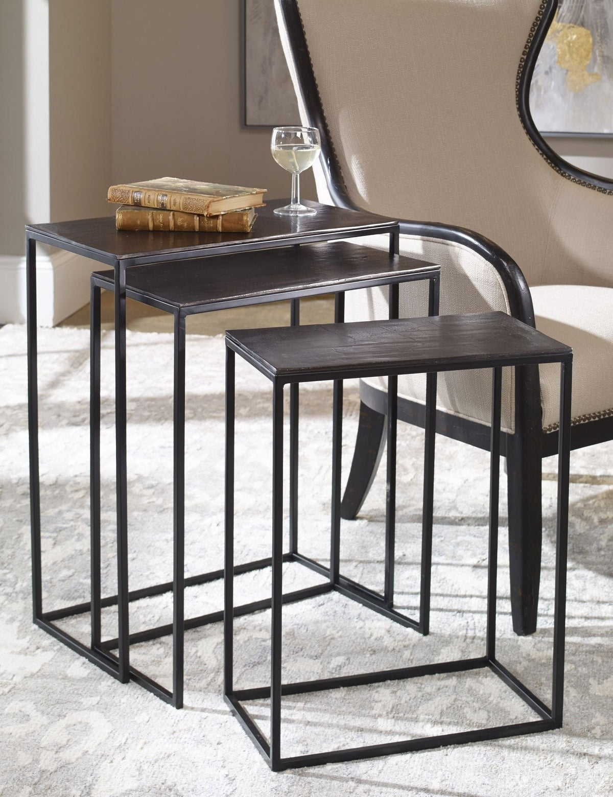  The Loletta black nesting tables are partially nested under each other, sitting in a living room next to a black wooden framed accent chair atop a gray patterned rug