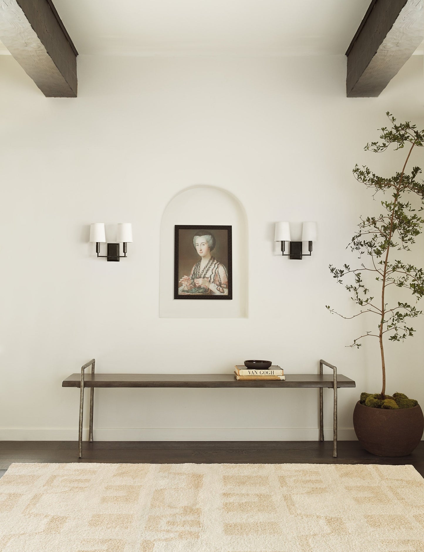 #color::dark-bronze | The Charlie dark bronze double sconce with parchment shades and angular arms is mounted in an entry way with a natural-toned plush rug, a portrait wall art, and long metal bench