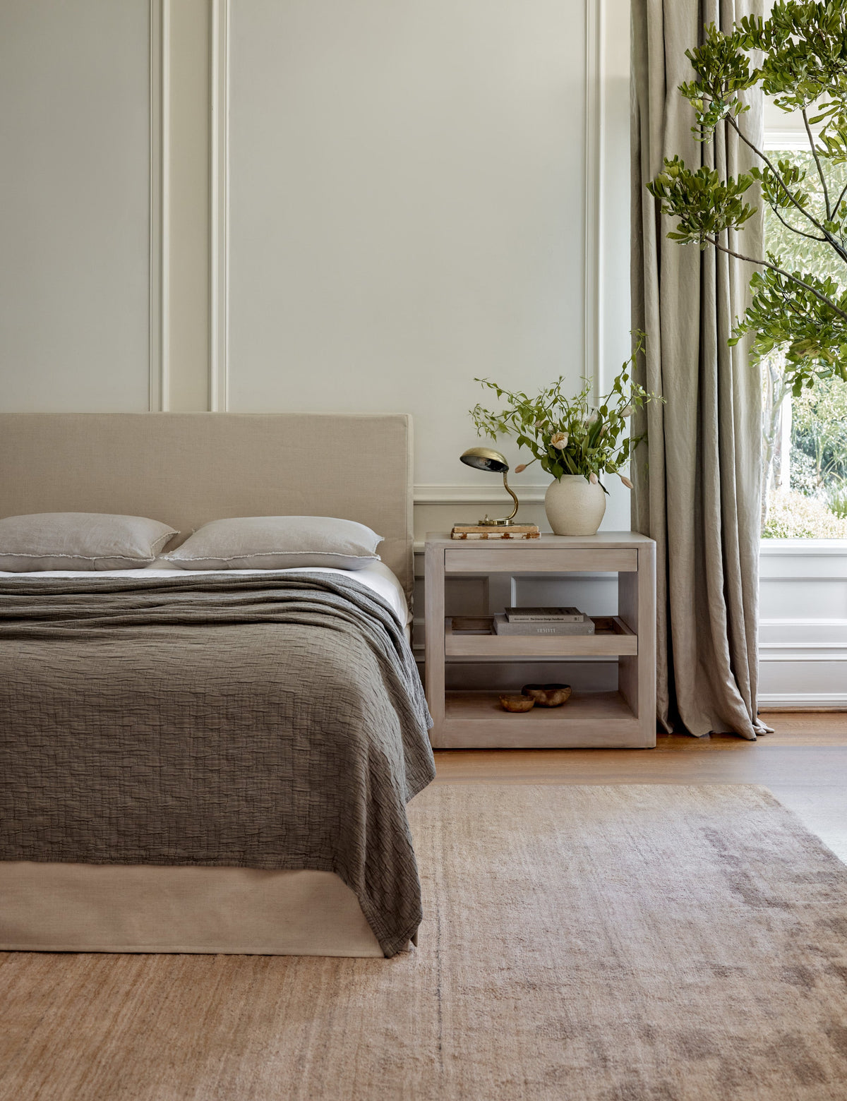 | A dark painted bedroom has the Arabel light wood open nightstand with two pull-out drawers sits next to an ivory upholstered bedframe with ivory grid bedding atop an ivory area rug with tan geometric border. 