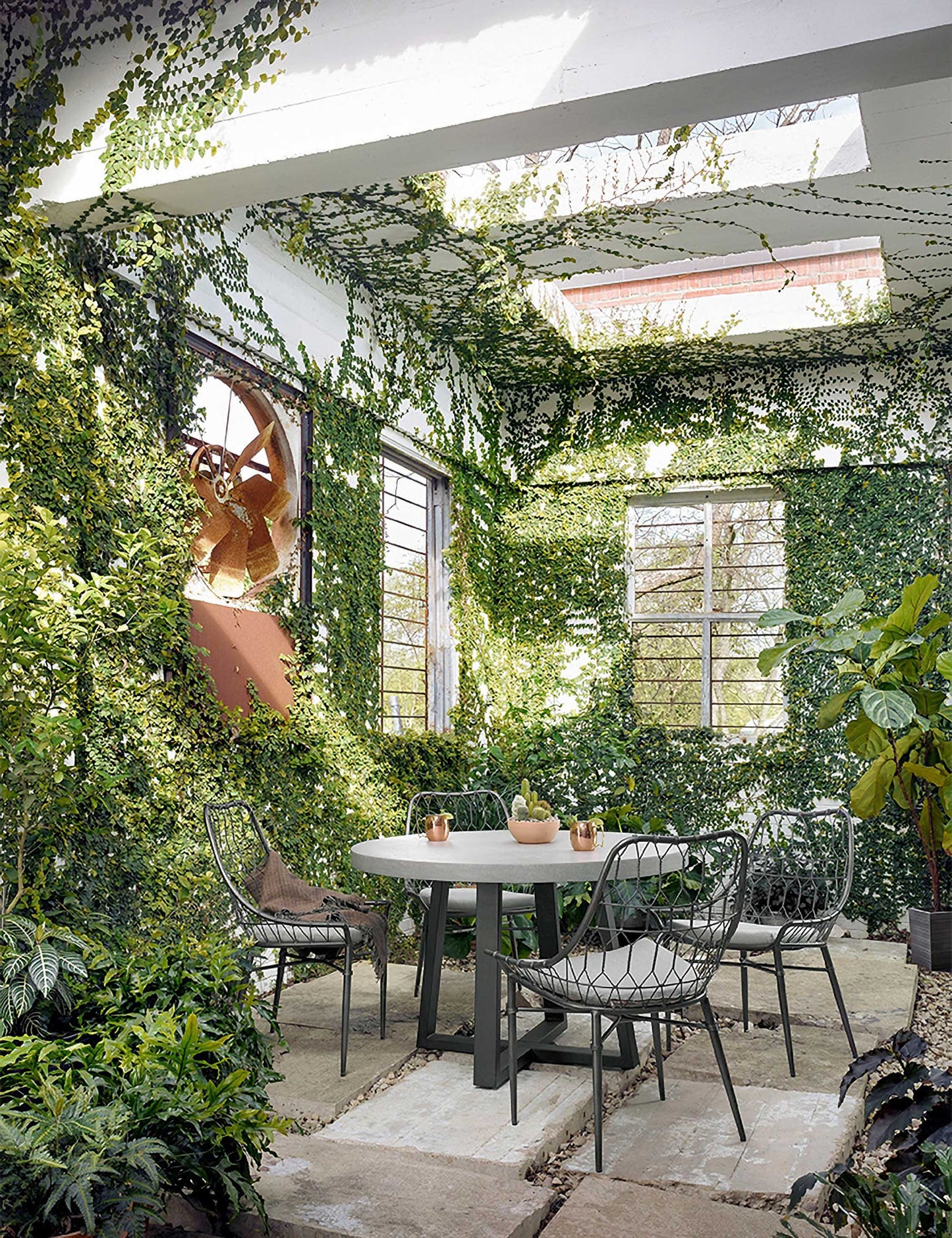 | The Agatha indoor and outdoor dining table with lavastone top and black iron base sits in a covered outdoor space with ivy-covered walls and black woven dining chairs 