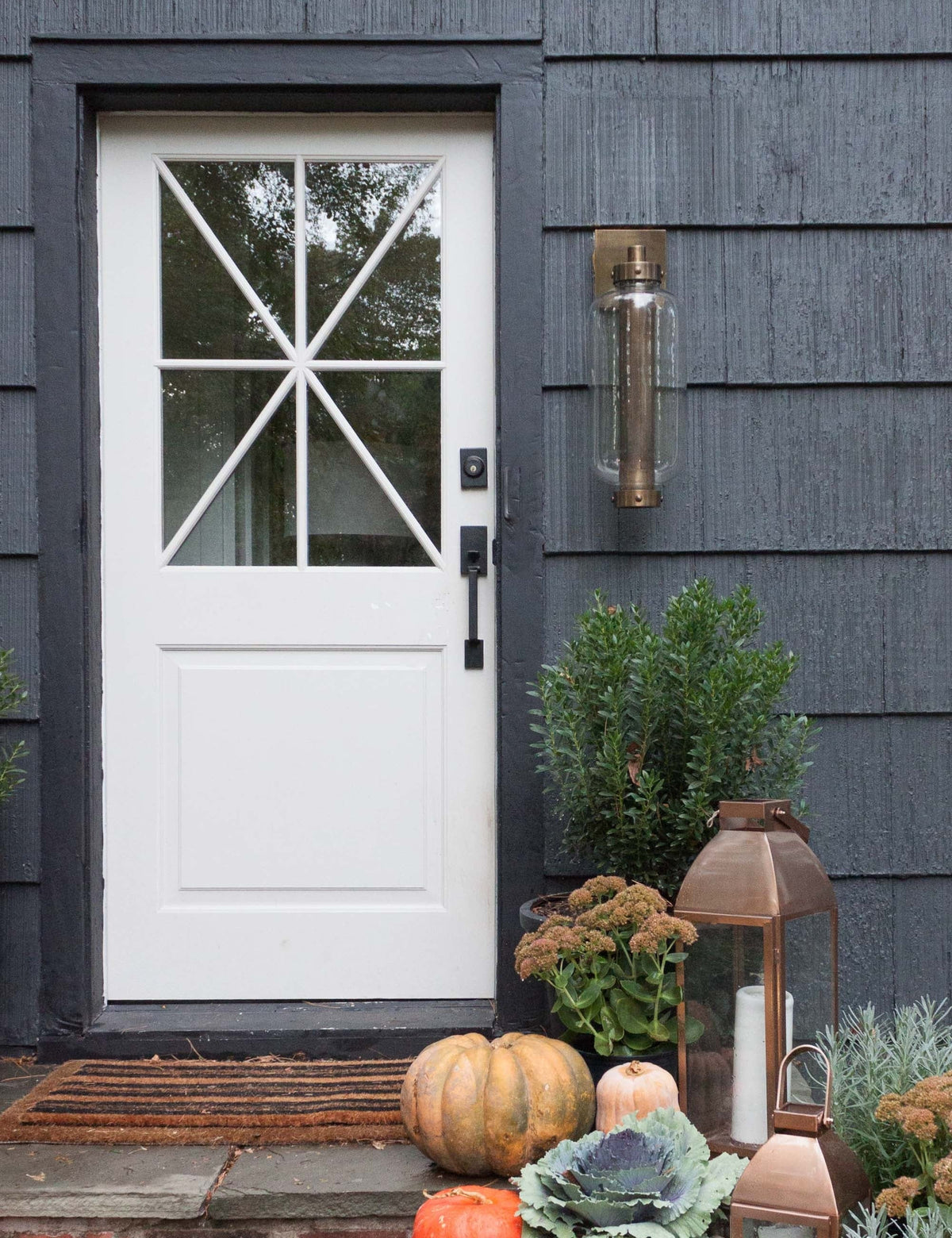| The Romelia indoor and outdoor sconce with metal mesh encased bulb and brass hardware hangs out the exterior of a black paneled home to the right of a white door