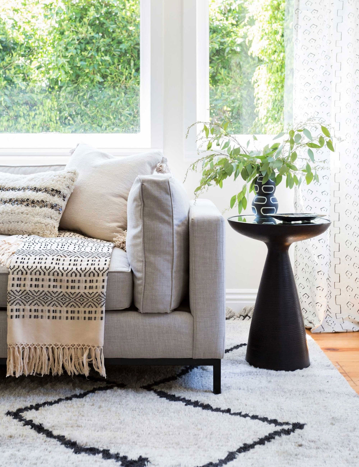 | The Bridget bronze pedestal side table with mirror ash glass top sits in a living room with a gray linen sofa, a plush patterned rug, and a geometric throw blanket