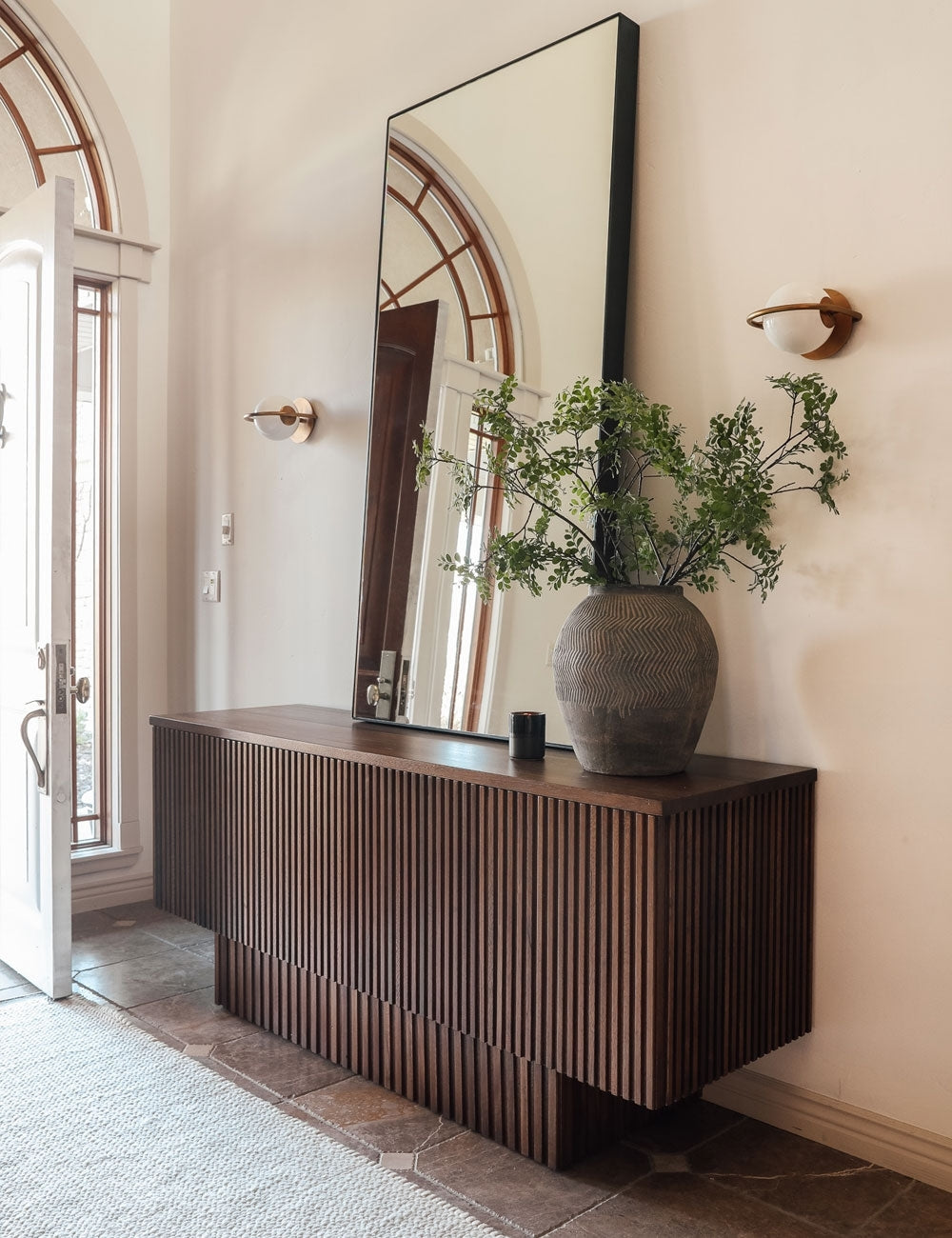 | The Evangeline diffused white glass orb sconces with brass frames sit in an entryway with a striped dark wooden sideboard and a tall rectangular mirror between them