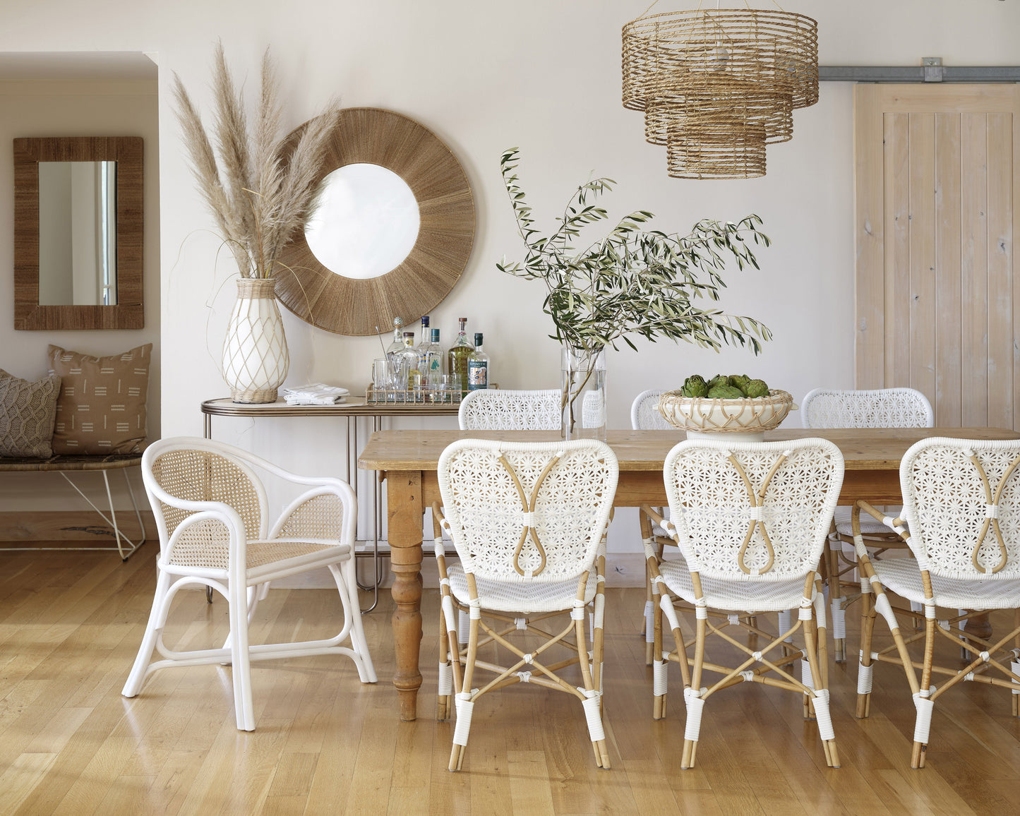 | The Minna three-tiered jute chandelier sits in a dining room with white woven dining chairs, a wooden dining table, and a jute framed circular mirror