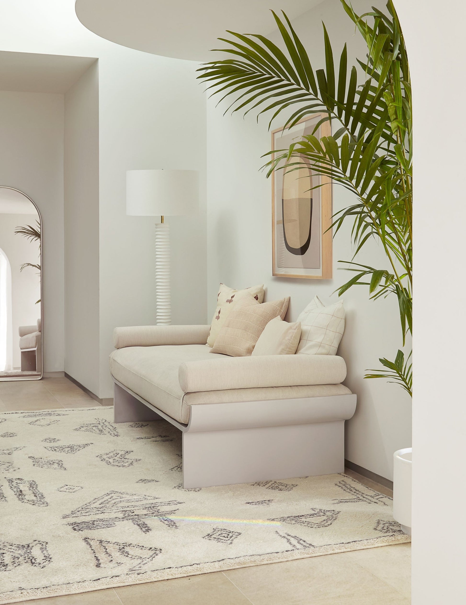 | The Nabu white metal floor lamp with ribbed base and gold accents by Regina Andrew sits in the corner of an entry way with a plush geometric rug, a beige linen sofa, and a natural toned wall art