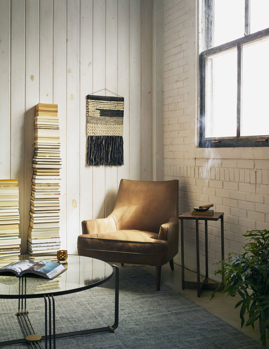 | The Ilona Leather Accent Chair sits in the corner of a living room next to a round coffee table and two high stacks of books