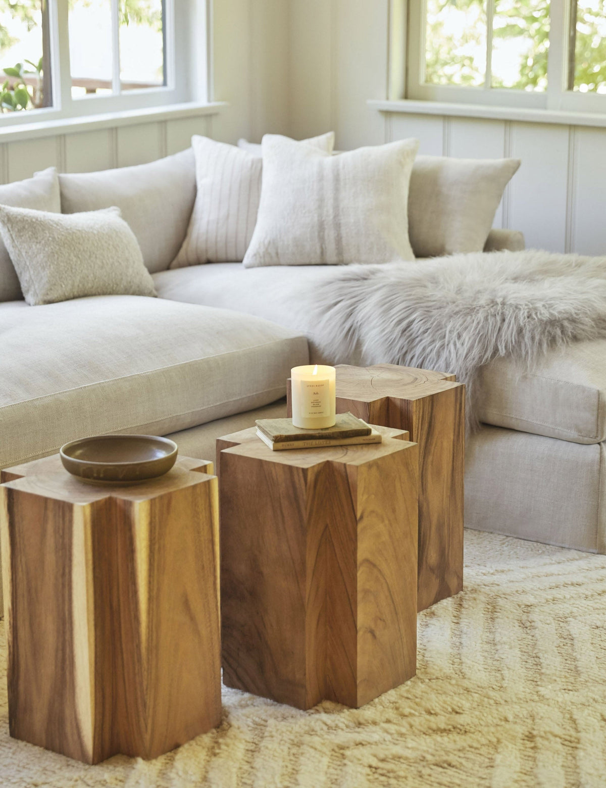 | Three Lolani plus shaped wooden side tables sit in a warm living room with a linen sectional, a gray plush sheepskin throw, and a white chevron patterned rug.