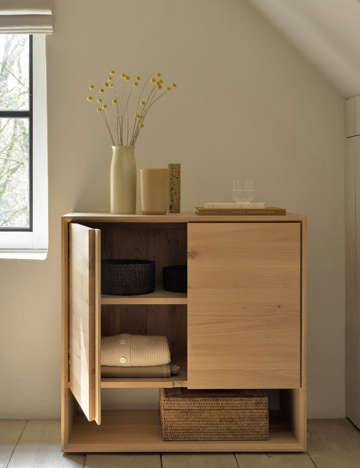 | The Lark 2-Door Sideboard with one door open sits in an attic space with a vase and book atop it