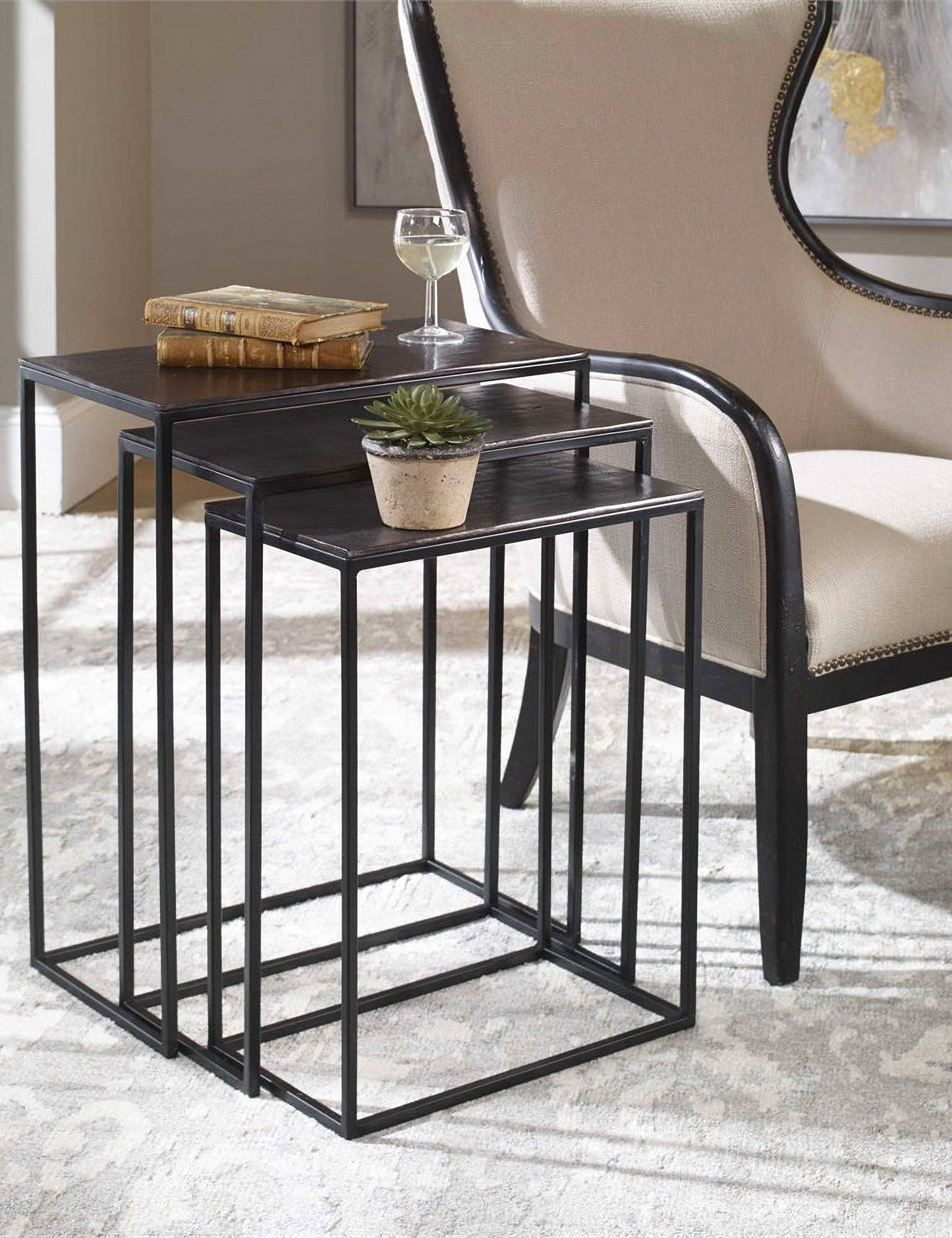| The Loletta black nesting tables are partially nested under each other, sitting in a living room next to a black wooden framed accent chair atop a gray patterned rug