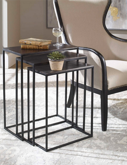 | The Loletta black nesting tables are partially nested under each other, sitting in a living room next to a black wooden framed accent chair atop a gray patterned rug