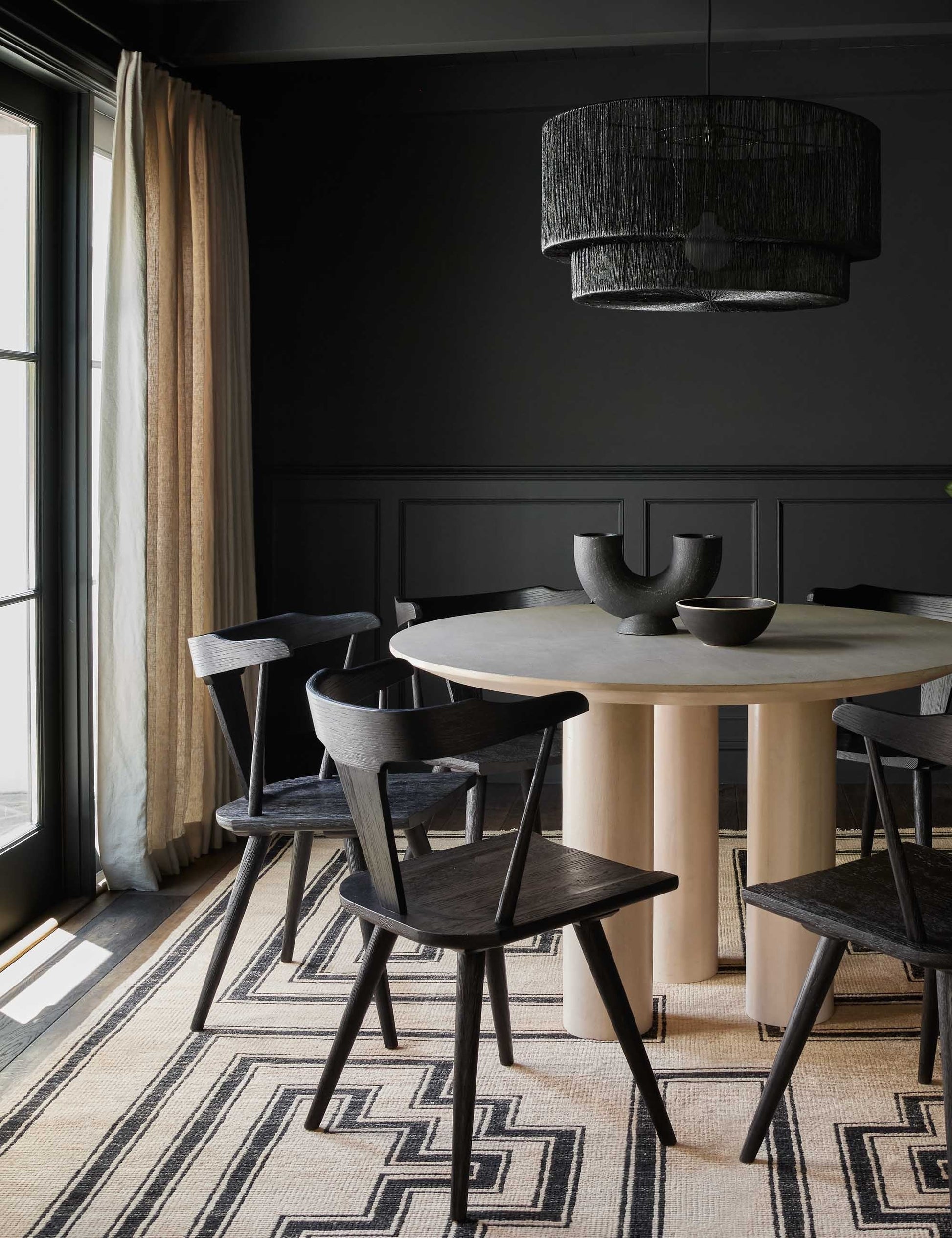 | The Damien half-circle sculpture vase by Ateriors with matte black glaze finish sits in a dark dining room with black accented walls, black dining chairs, and a black and beige geometric rug