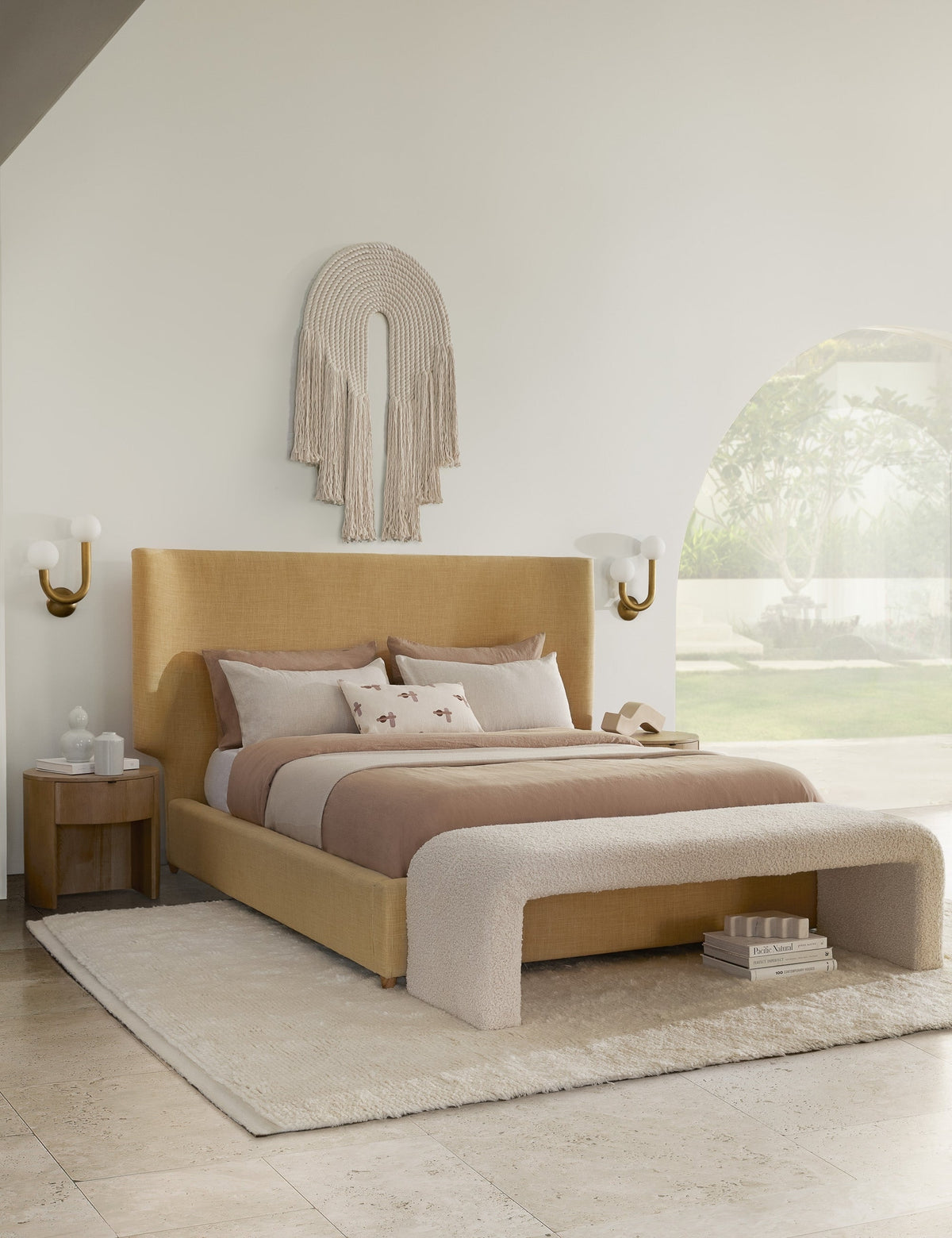 | The Kono round solid oak nightstand with one drawer has white accessories on it and sits next to a gold fabric upholstered bed with tan and ivory bedding, a curved bench, and an arched woven wall hanging above the bed.